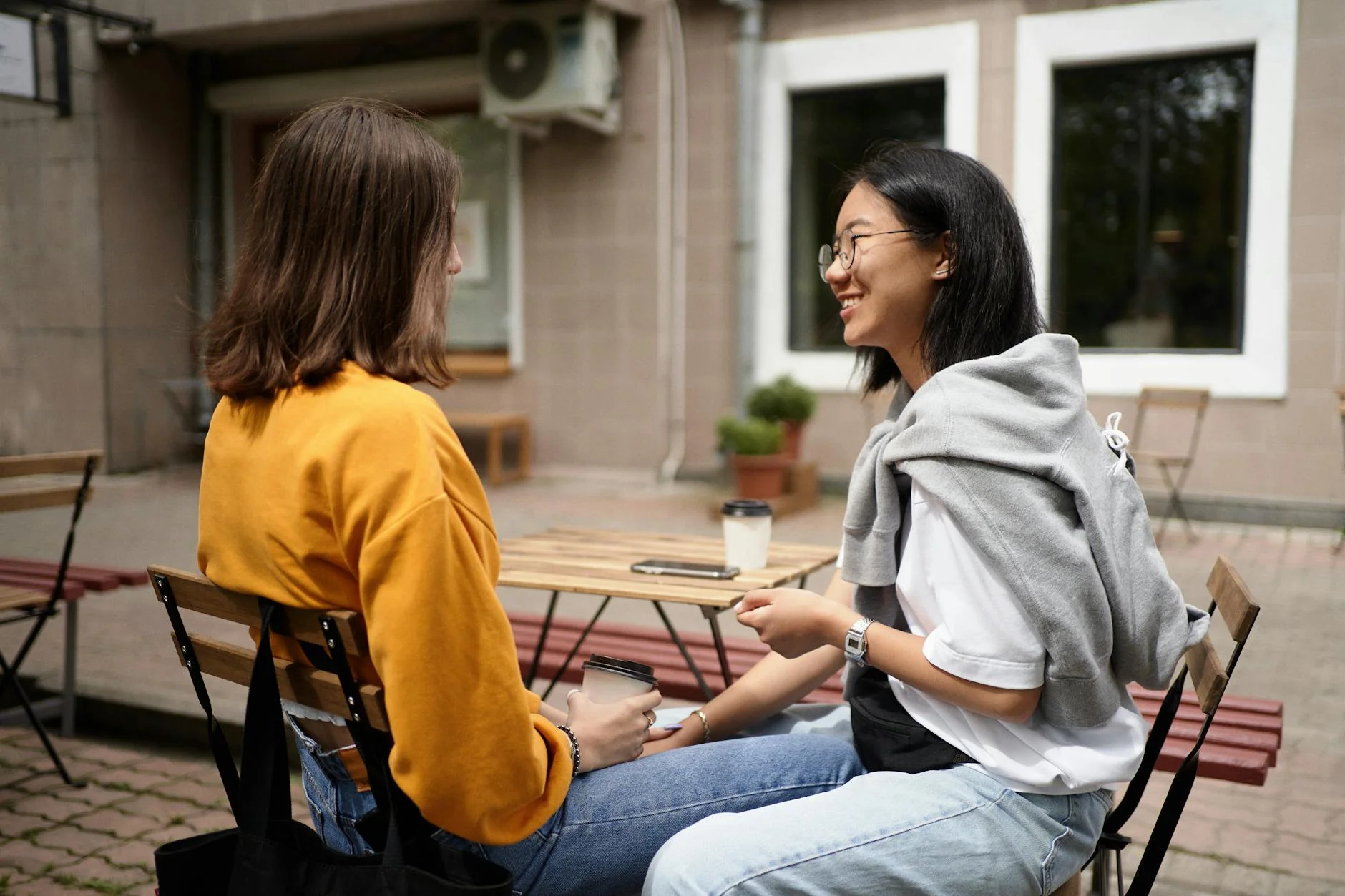 Dos mujeres conversando en una mesa de cafetería luminosa con tazas y libretas abiertas.